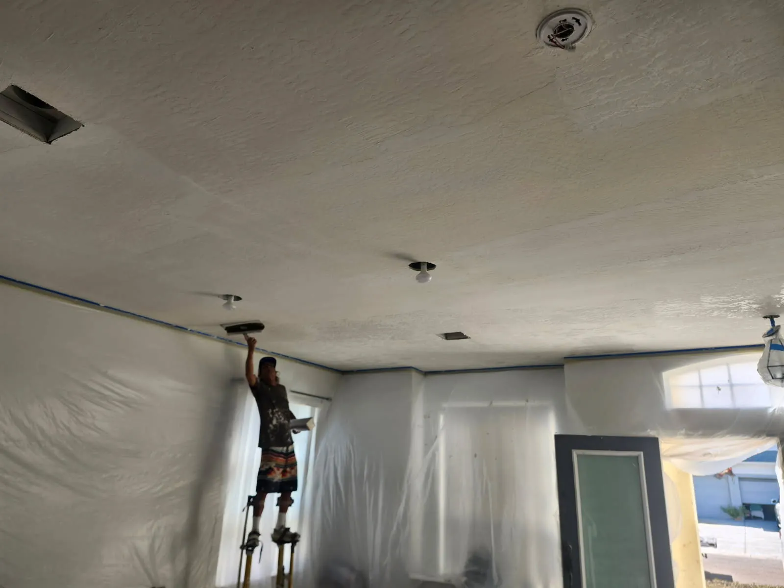Bill Roebuck, owner of WG Roebuck Drywall, on stilts working a knockdown texture across a living room ceiling, with the walls, windows and doors masked in plastic